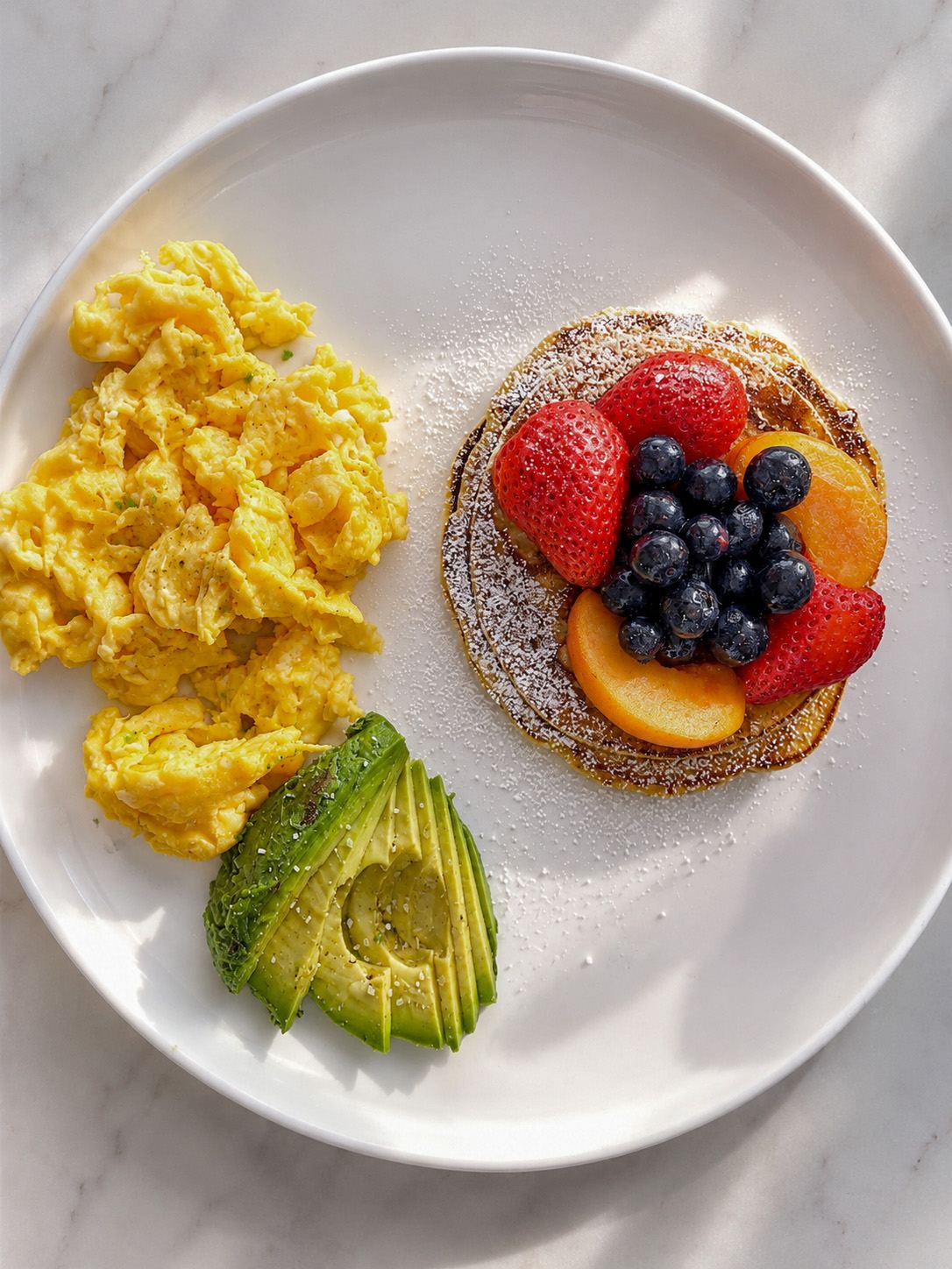 Overhead plate of scrambled eggs, avocado, and a berry-topped pancake on marble