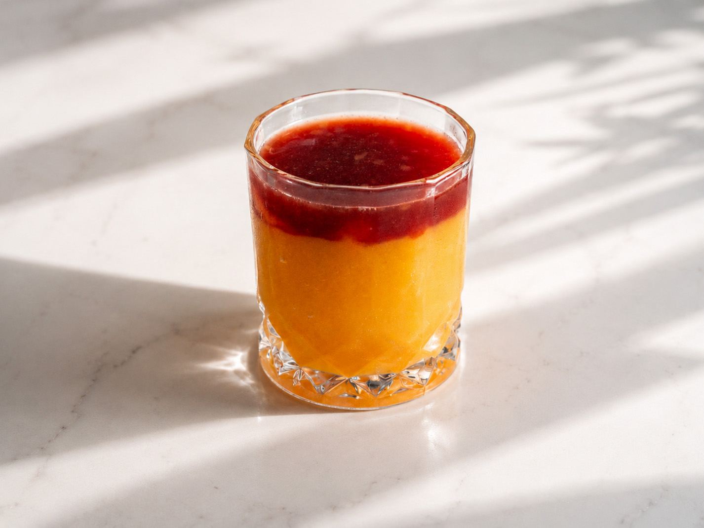 Layered peach and berry drink in a cut-glass tumbler in raking sunlight